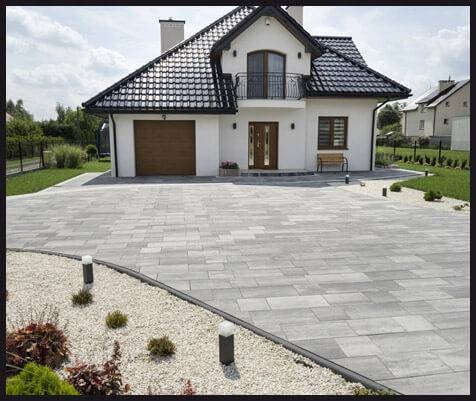 Szeroki podjazd z szarej kostki brukowej prowadzący do białego domu z brązową bramą garażową i czarnym dachem, obrzeża z białego żwiru i niskie lampy oświetleniowe.