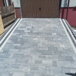 Ułożona szara kostka brukowa z obramowaniem z jasnego grysu i ciemnoszarej kostki, prowadząca do brązowej bramy garażowej, widoczna łopata oparta o ścianę budynku.