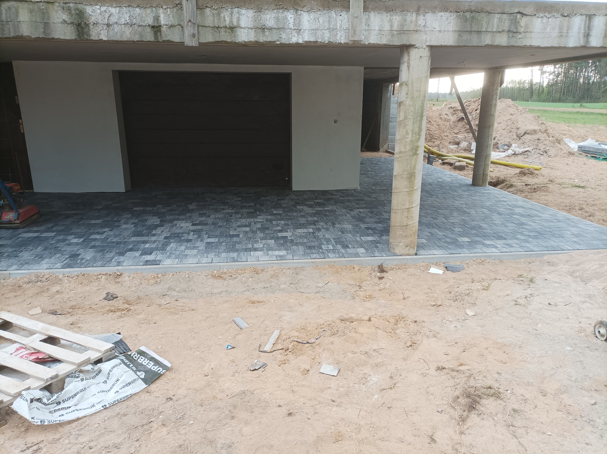 Nowa, szara kostka brukowa ułożona przed garażem w surowym stanie, widoczny paleta i resztki materiałów budowlanych na piaszczystym podłożu, fragment niedokończonego budynku w tle.