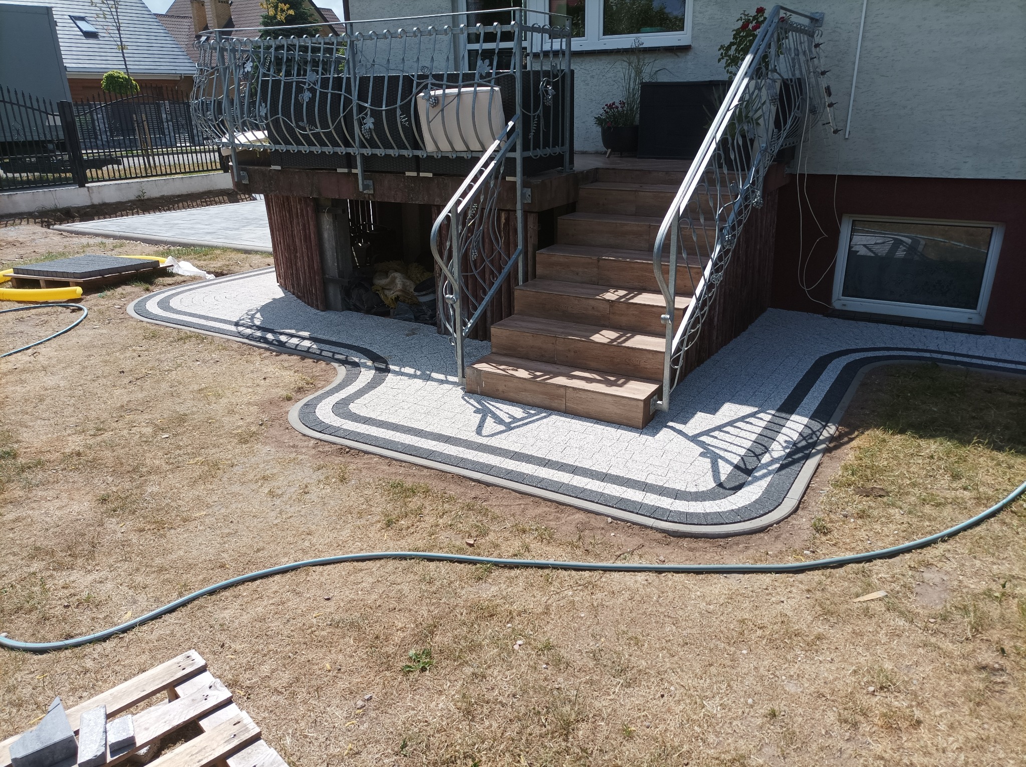 Ułożona kostka brukowa w jasnych odcieniach z ciemnoszarą obwódką tworząca ścieżkę do drewnianych schodów z metalową balustradą, widoczne elementy ogrodu i domu.