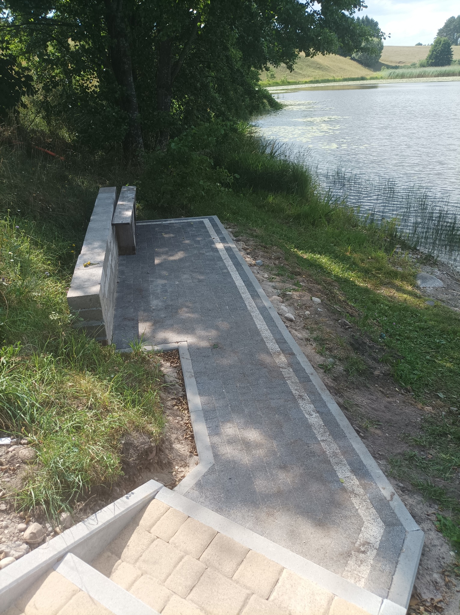 Ścieżka z kostki brukowej prowadząca do jeziora, z betonowymi schodami i ławką z bloków betonowych w tle, otoczona zielenią i częściowo niedokończonym terenem.