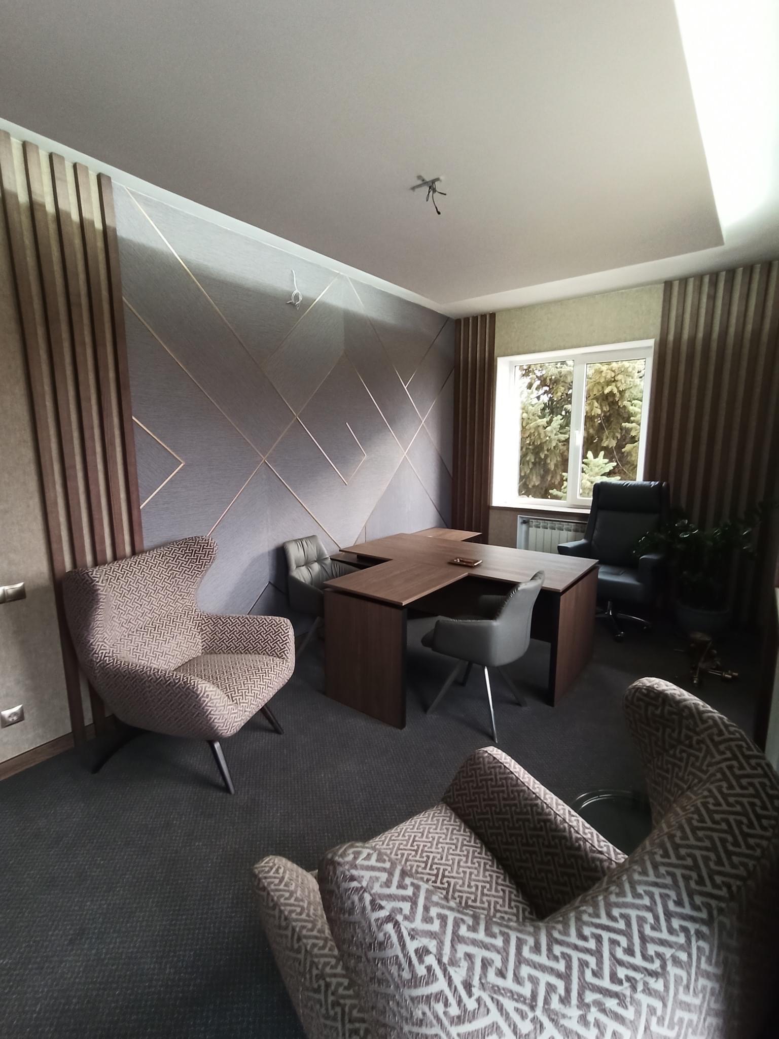 Biuro z drewnianym biurkiem w kształcie litery L, fotelami w geometryczne wzory, szarą ścianą z dekoracyjnymi listwami i oknem z widokiem na zieleń.