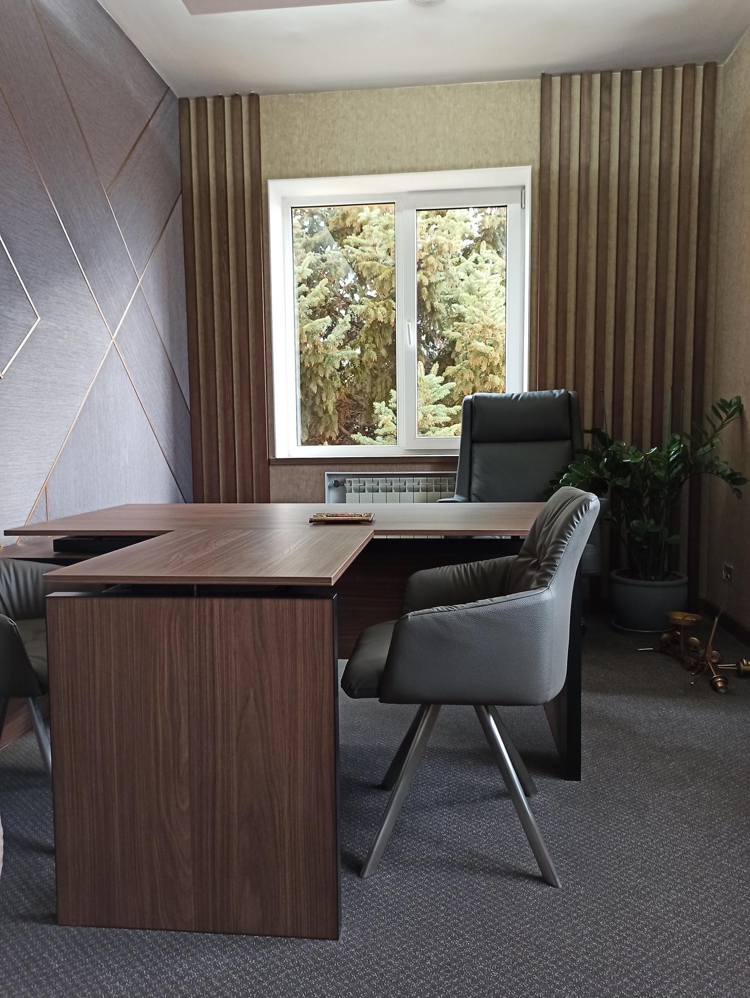 Nowoczesne biuro z biurkiem w kształcie litery L z ciemnego drewna, krzesłami na metalowych nogach i dużym oknem z widokiem na drzewa, dekoracyjne panele ścienne w odcieniach szarości i brązu.