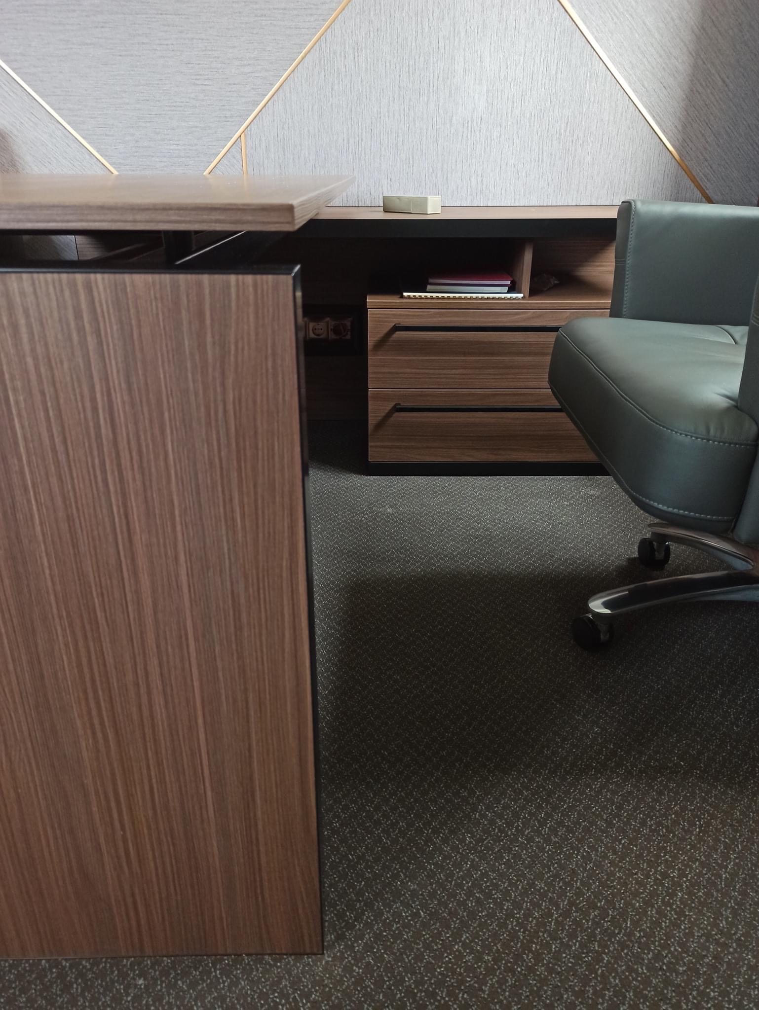 Nowoczesne biuro z biurkiem z drewnianym wykończeniem, szarą tapetą w geometryczne wzory i ergonomicznym krzesłem na kółkach.