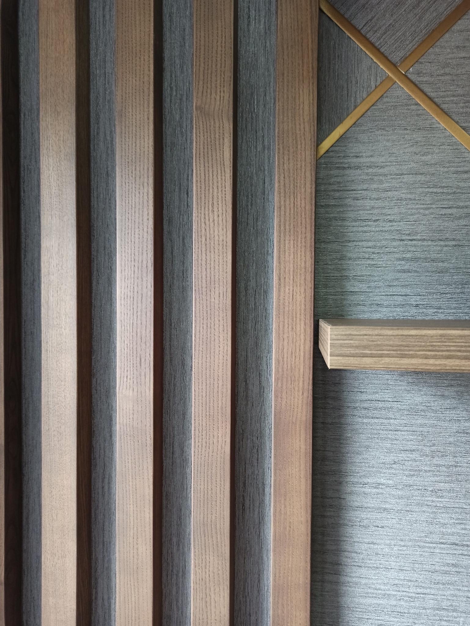 Drewniane lamele ścienne w odcieniach szarości i brązu, przylegające do szarej ściany z dekoracyjnymi, złotymi listwami i małą drewnianą półką.
