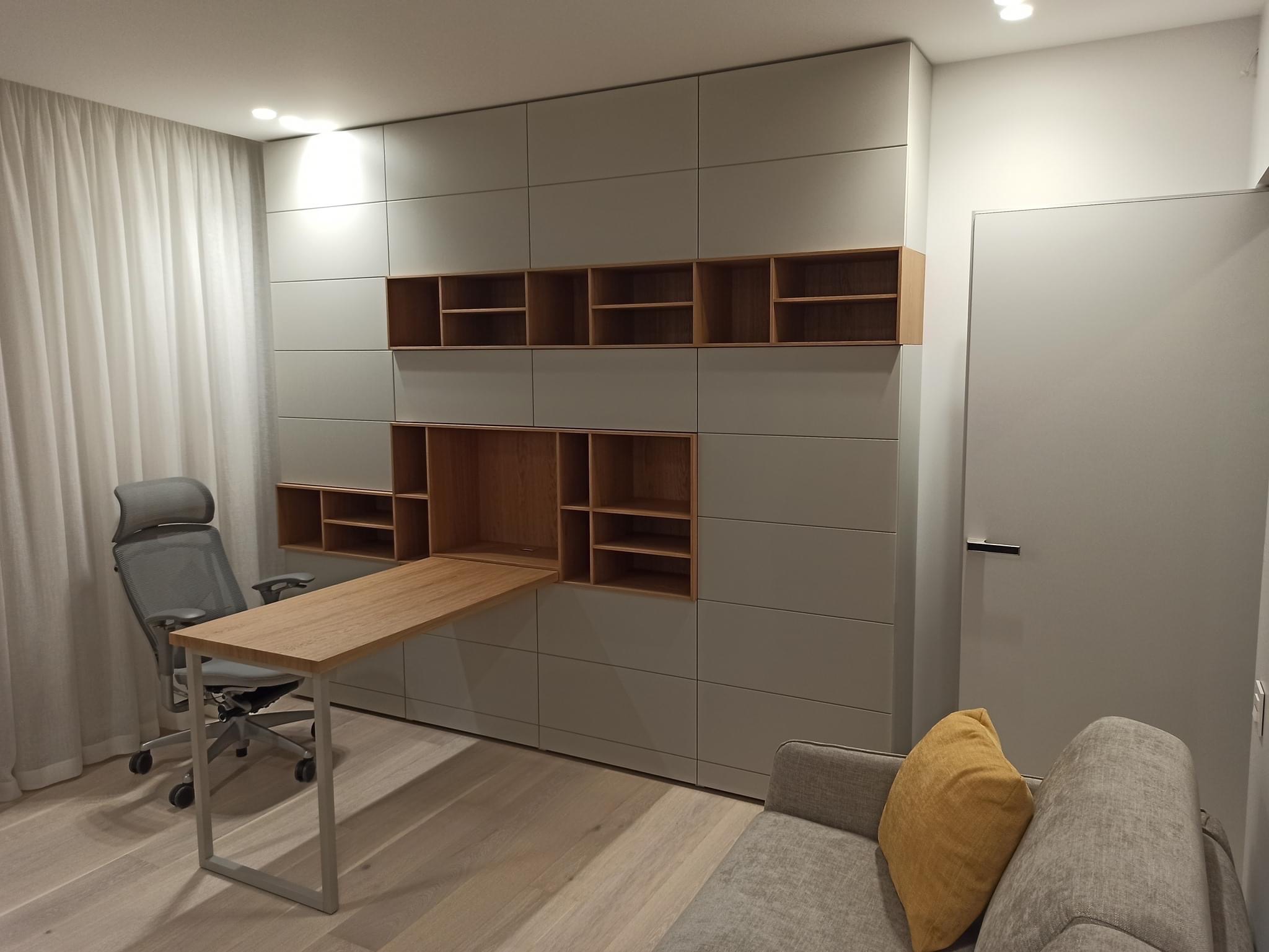 Szara zabudowa ścienna z drewnianymi półkami i rozkładanym blatem biurka, uzupełniona o ergonomiczne krzesło biurowe i fragment szarej sofy z żółtą poduszką.