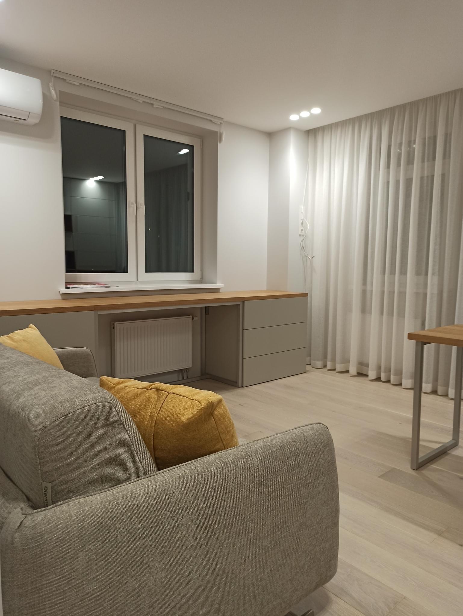 Jasnoszary pokój z biurkiem pod oknem, szarą sofą z żółtymi poduszkami i białymi zasłonami, oświetlony trzema punktowymi lampami sufitowymi.