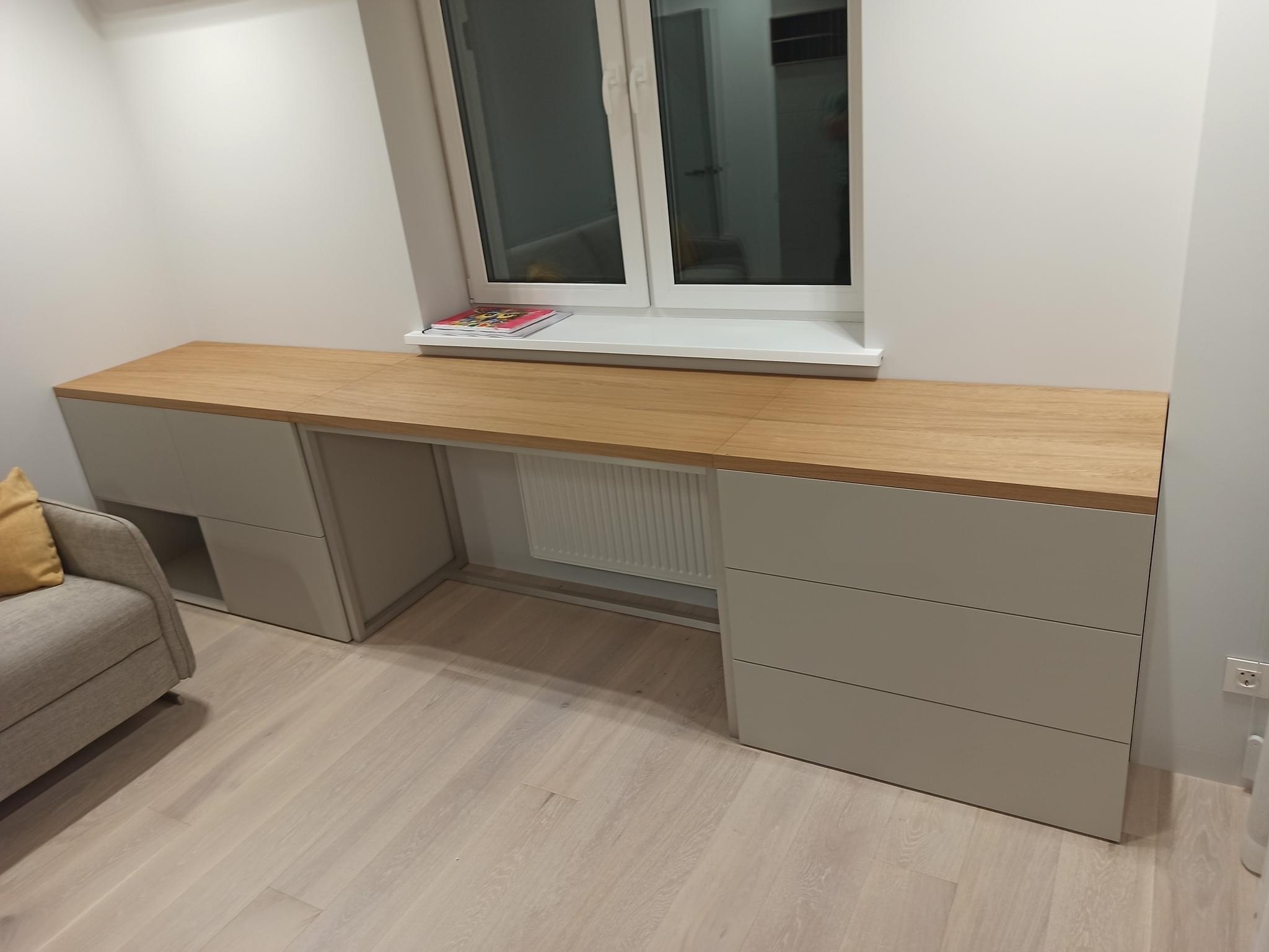 Biurko z drewnianym blatem i szarymi szafkami w zabudowie wnęki okiennej w Bytomiu, widoczny fragment sofy z żółtą poduszką.