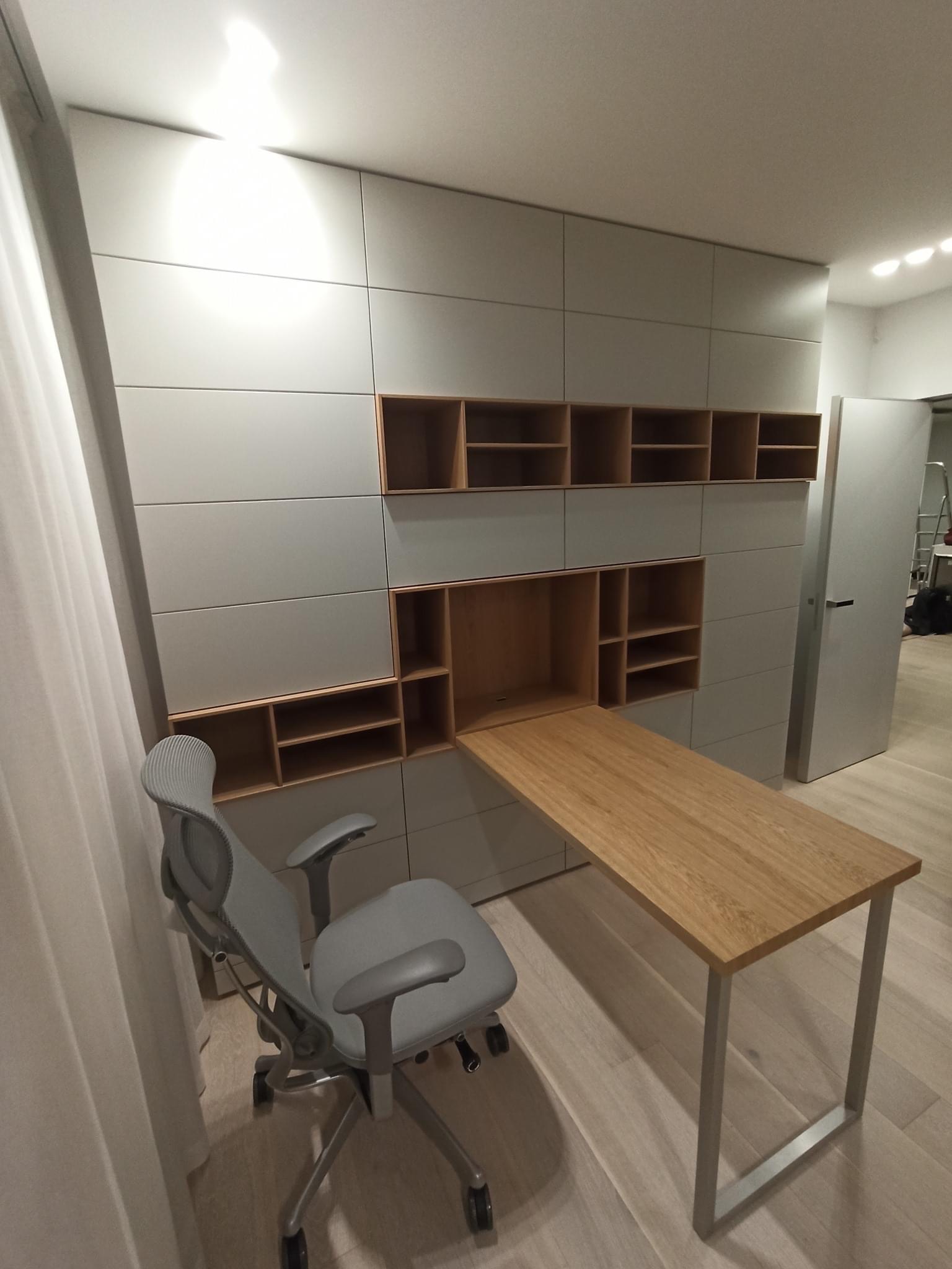 Szara ściana zabudowana szafami z drewnianymi wnękami i rozkładanym blatem biurka, szare krzesło biurowe na kółkach, jasna podłoga.