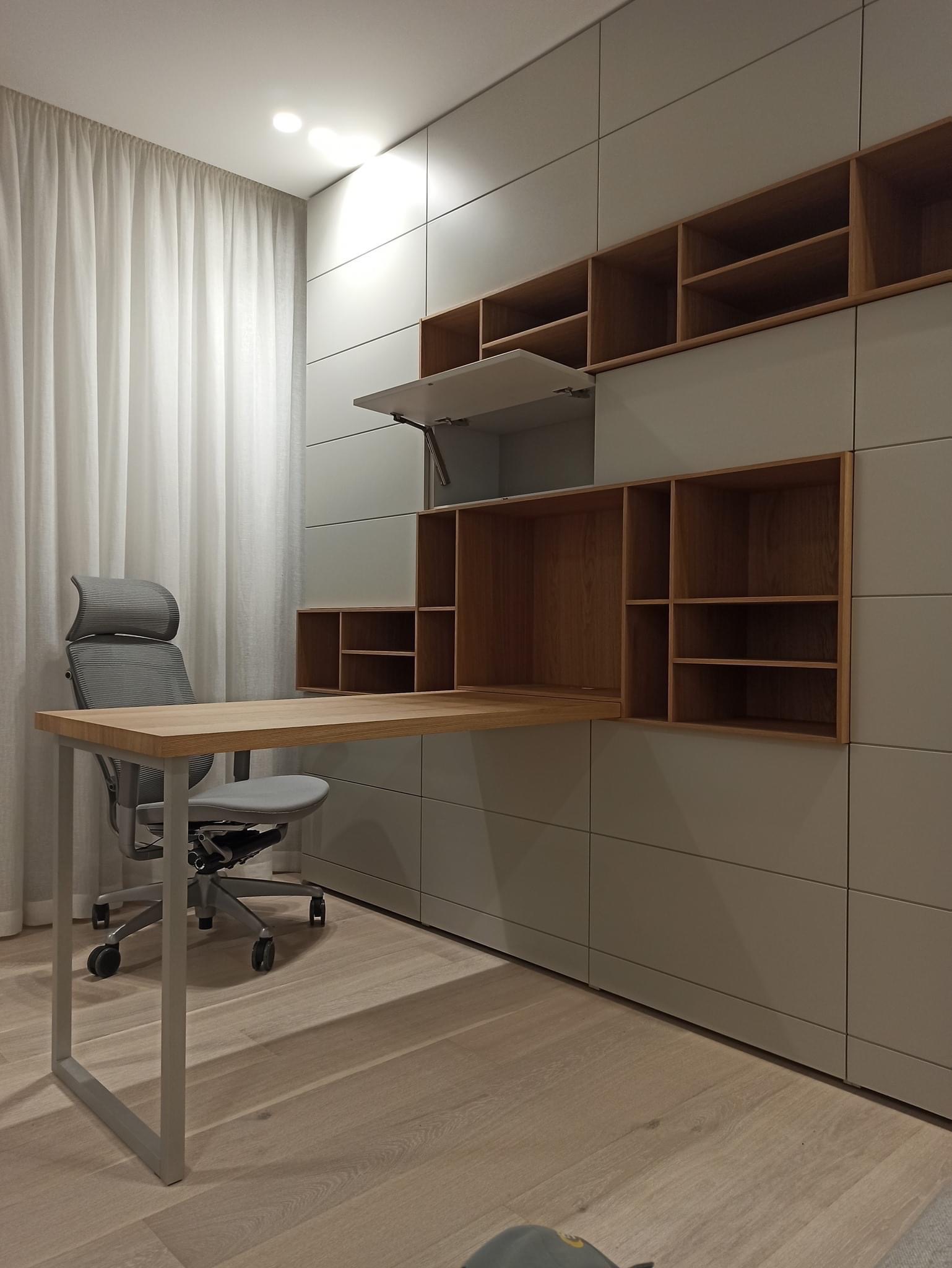 Nowoczesna aranżacja biura domowego z szarymi szafami w zabudowie, drewnianymi półkami i blatem biurka, oraz ergonomicznym krzesłem biurowym na kółkach.