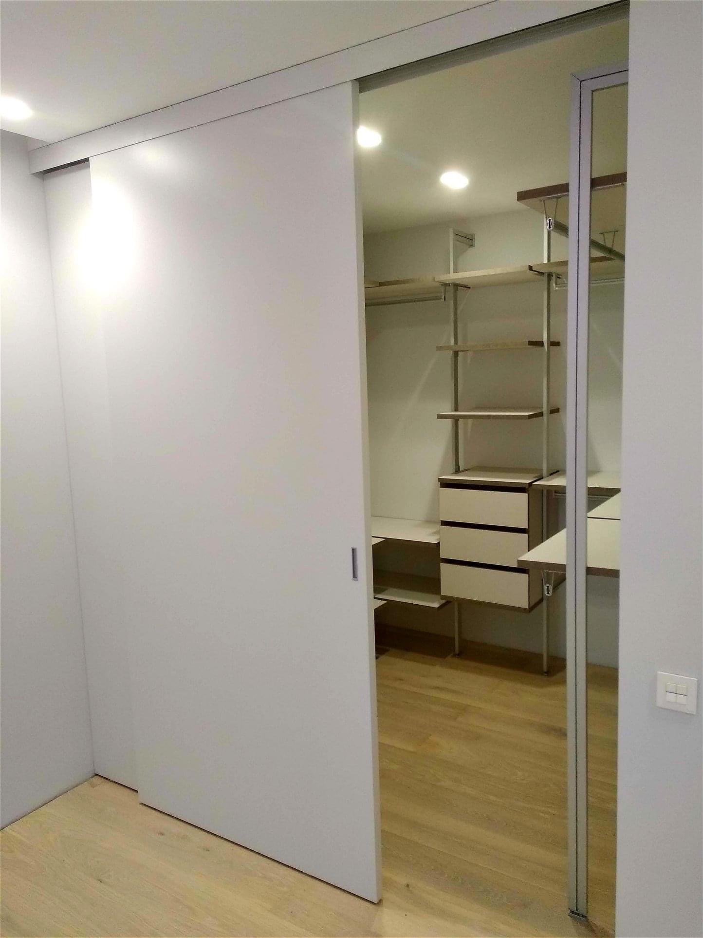 Biała szafa wnękowa z przesuwnymi drzwiami, widoczna z boku, z wnętrzem wyposażonym w półki i szuflady, oświetlona punktowymi lampami sufitowymi, na jasnej drewnianej podłodze.