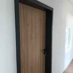 Zamknięte drzwi wewnętrzne z drewnopodobną okleiną i czarną ościeżnicą w białym korytarzu, widoczne klucze w zamku.