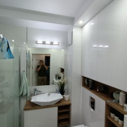 Lux-House - Nowoczesna łazienka z białymi płytkami, umywalką nablatową o geometrycznym kształcie, drewnianym blatem i szafkami, widoczne lustro z odbiciem osoby robiącej zdjęcie.