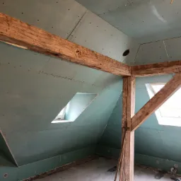 Poddasze z drewnianą konstrukcją, obłożone zielonymi płytami gipsowo-kartonowymi, widoczne okna dachowe i instalacje w podłodze.