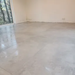 Jasnoszara, matowa podłoga z płytek ceramicznych w nowym, pustym pomieszczeniu z dużym oknem z widokiem na drzewa; widoczne gniazdka elektryczne przy ścianie.