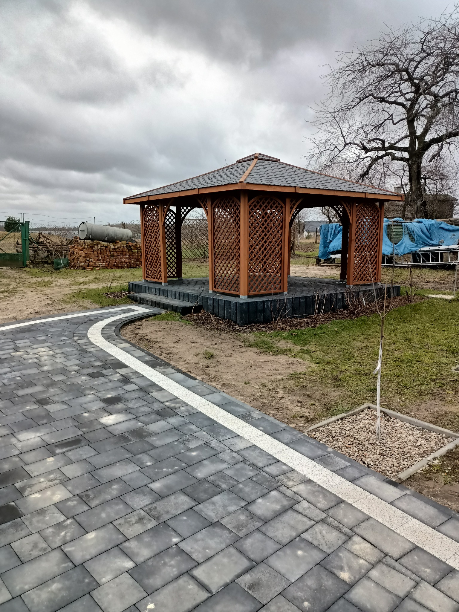Drewniana altana z ażurowymi ścianami i schodami, otoczona szarą kostką brukową z białym pasem, na tle pochmurnego nieba i drzew bez liści.