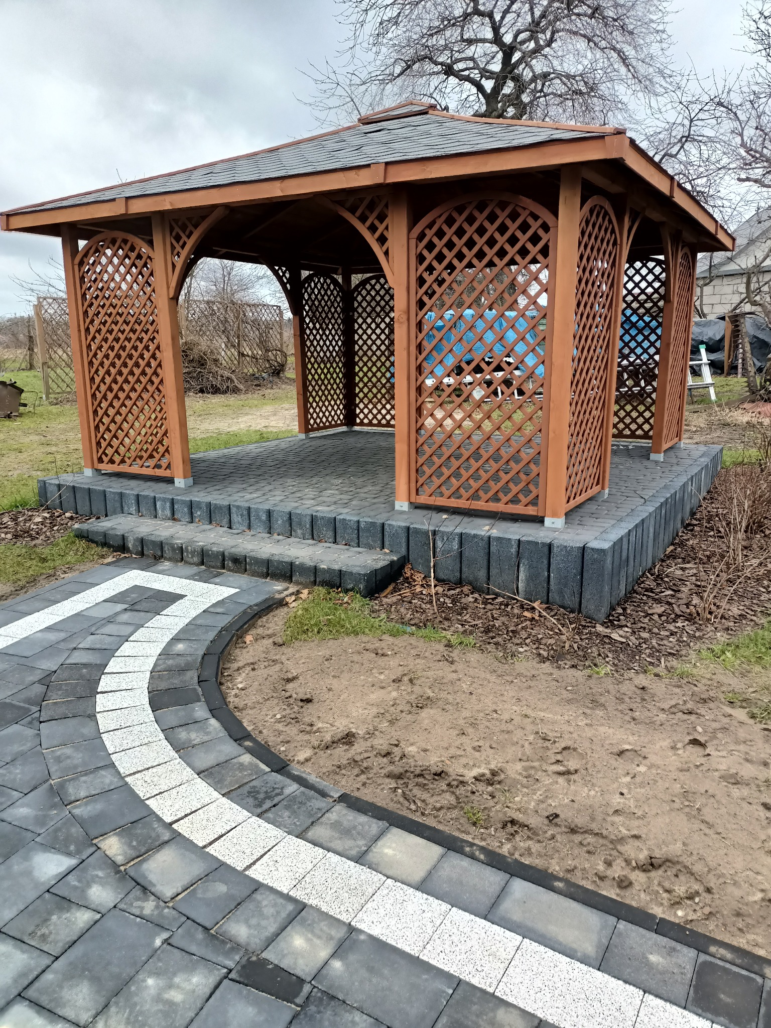 Drewniana altana z kratownicowymi ścianami, posadowiona na podwyższeniu z szarej kostki, połączona z ścieżką z kostki brukowej w odcieniach szarości z białym pasem, widok z perspektywy chodnika.