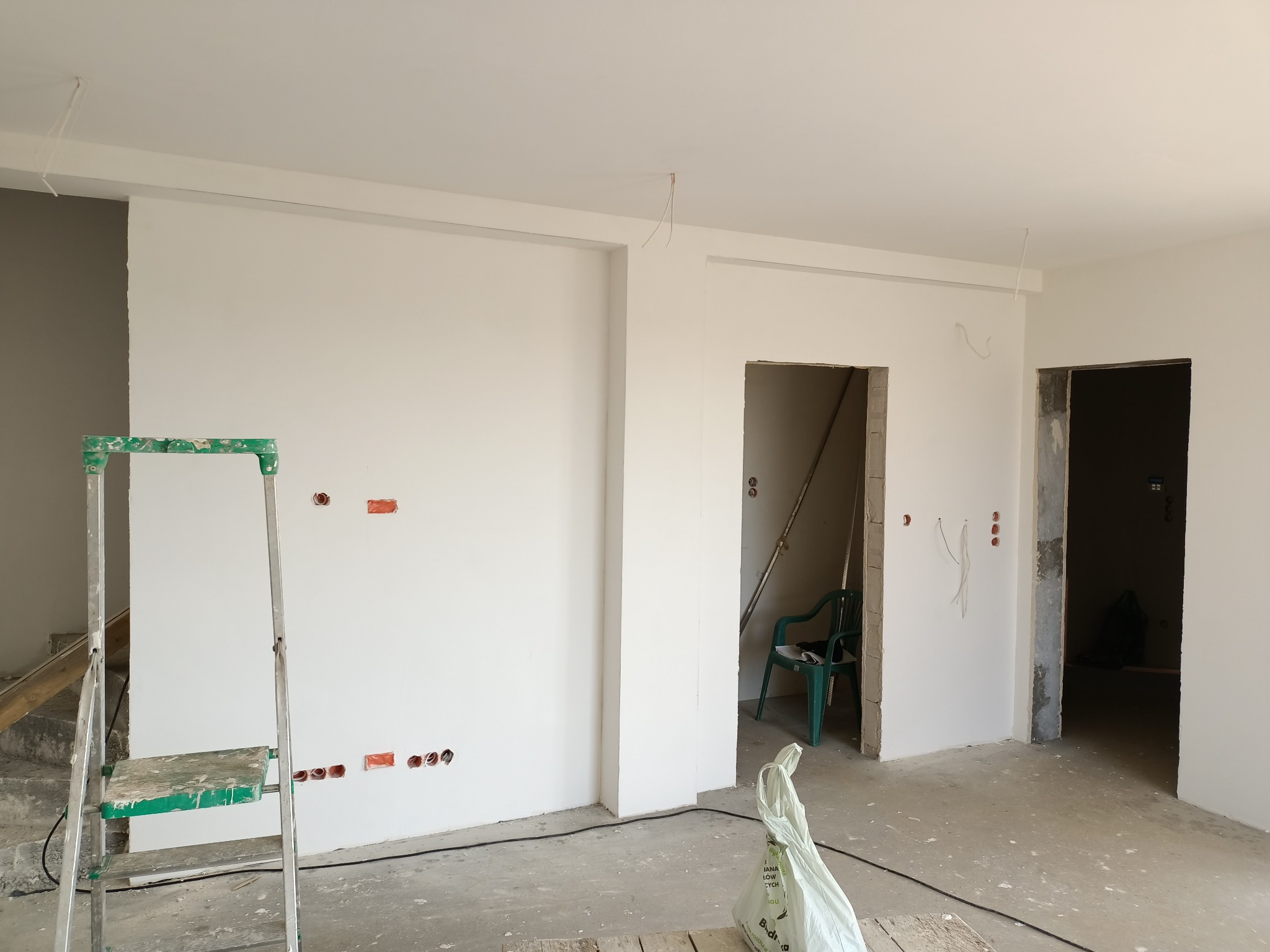 Wnętrze w trakcie prac wykończeniowych: białe ściany po szpachlowaniu i malowaniu, widoczne puszki elektryczne, drabina malarska z resztkami farby, surowe otwory drzwiowe i wystające kable.