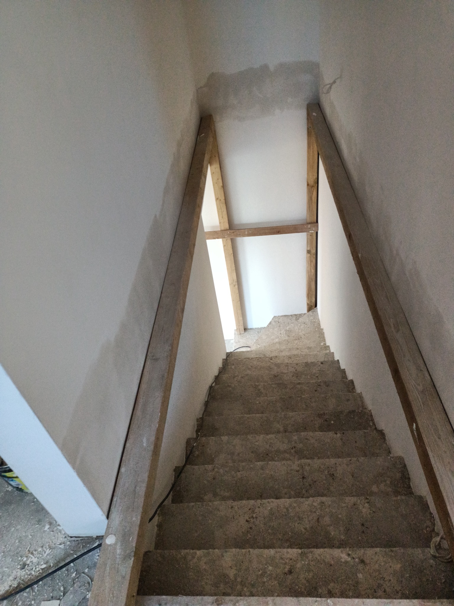 Surowe betonowe schody w trakcie remontu z drewnianą konstrukcją wspierającą, ściany częściowo pokryte świeżym tynkiem.