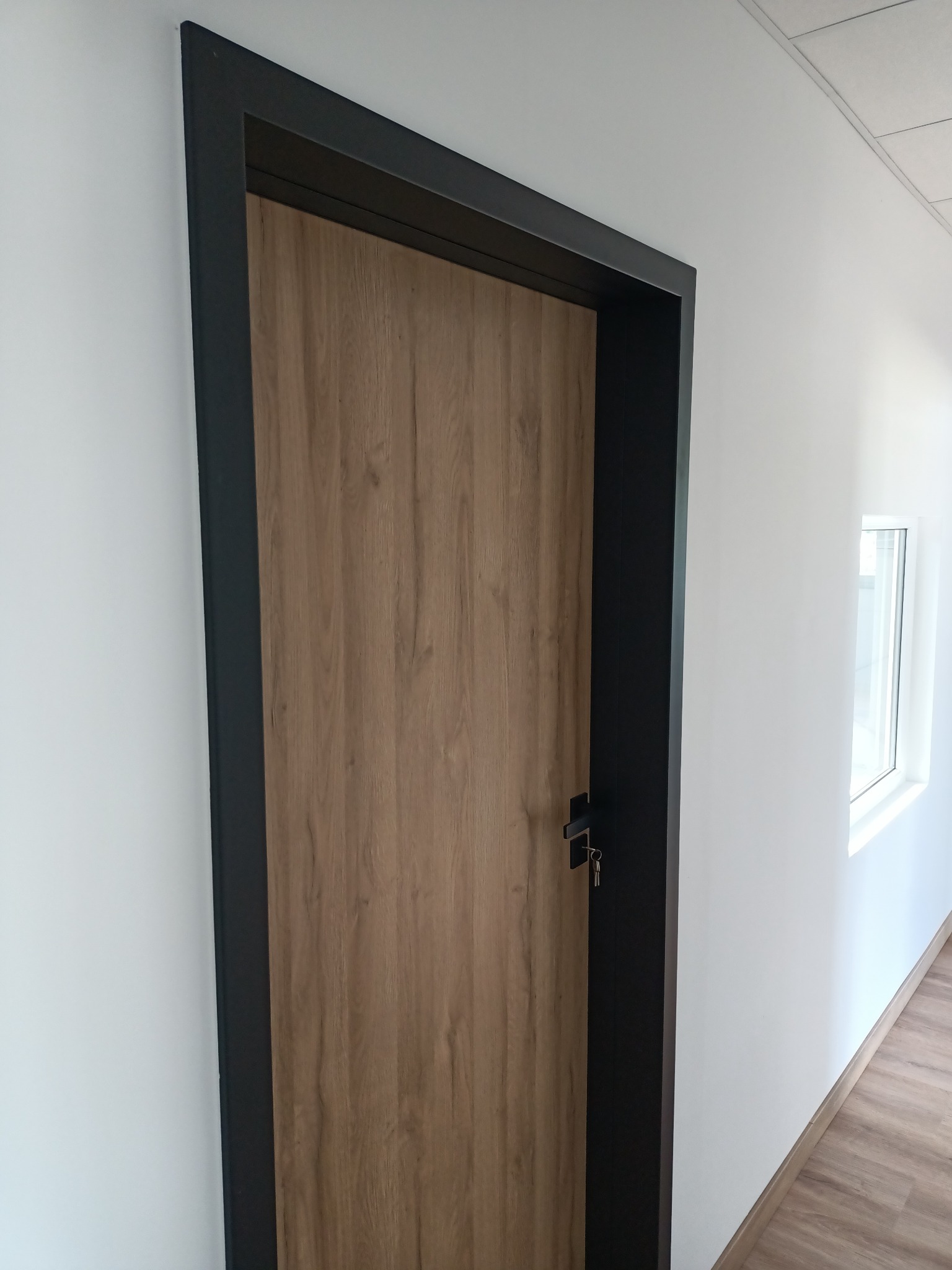 Zamknięte drzwi wewnętrzne z drewnopodobną okleiną i czarną ościeżnicą w białym korytarzu, widoczne klucze w zamku.