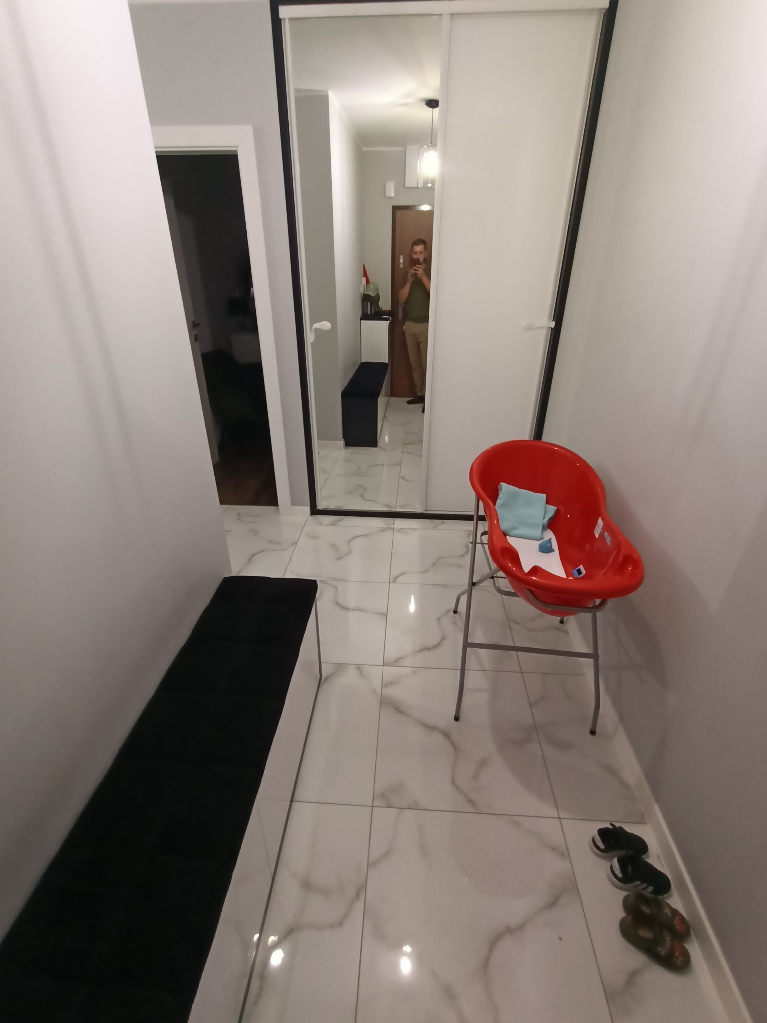 Przedpokój w Kościerzynie z białymi ścianami, marmurową podłogą, przesuwnymi drzwiami z lustrem, ławką z czarnym siedziskiem i czerwonym krzesełkiem dla dziecka.