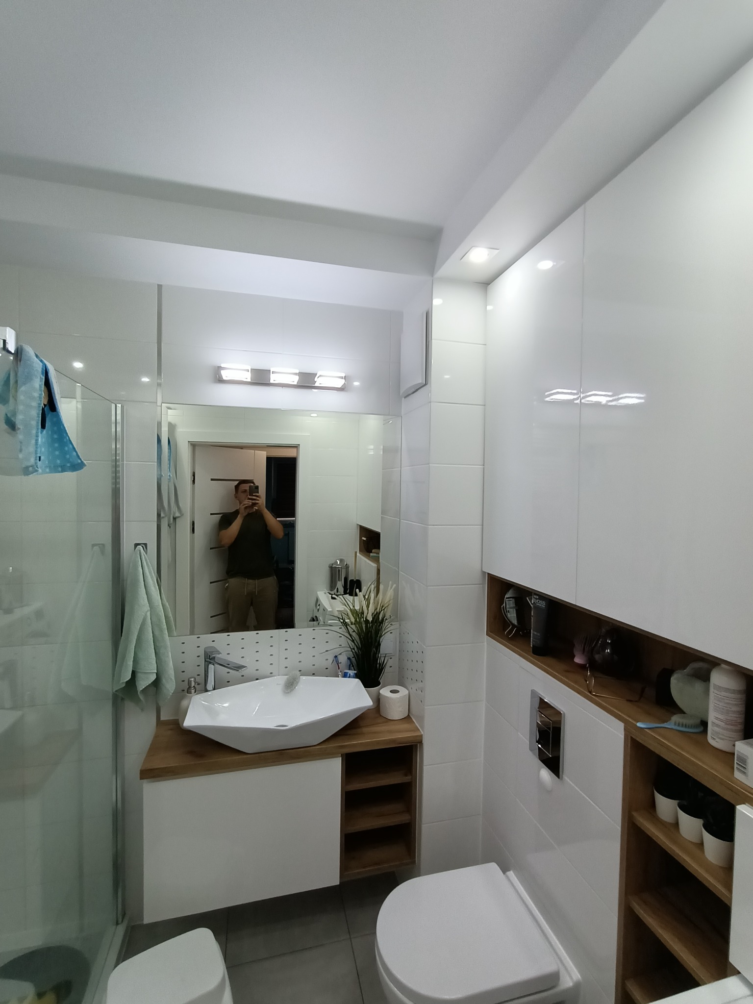 Nowoczesna łazienka z białymi płytkami, umywalką nablatową o geometrycznym kształcie, drewnianym blatem i szafkami, widoczne lustro z odbiciem osoby robiącej zdjęcie.