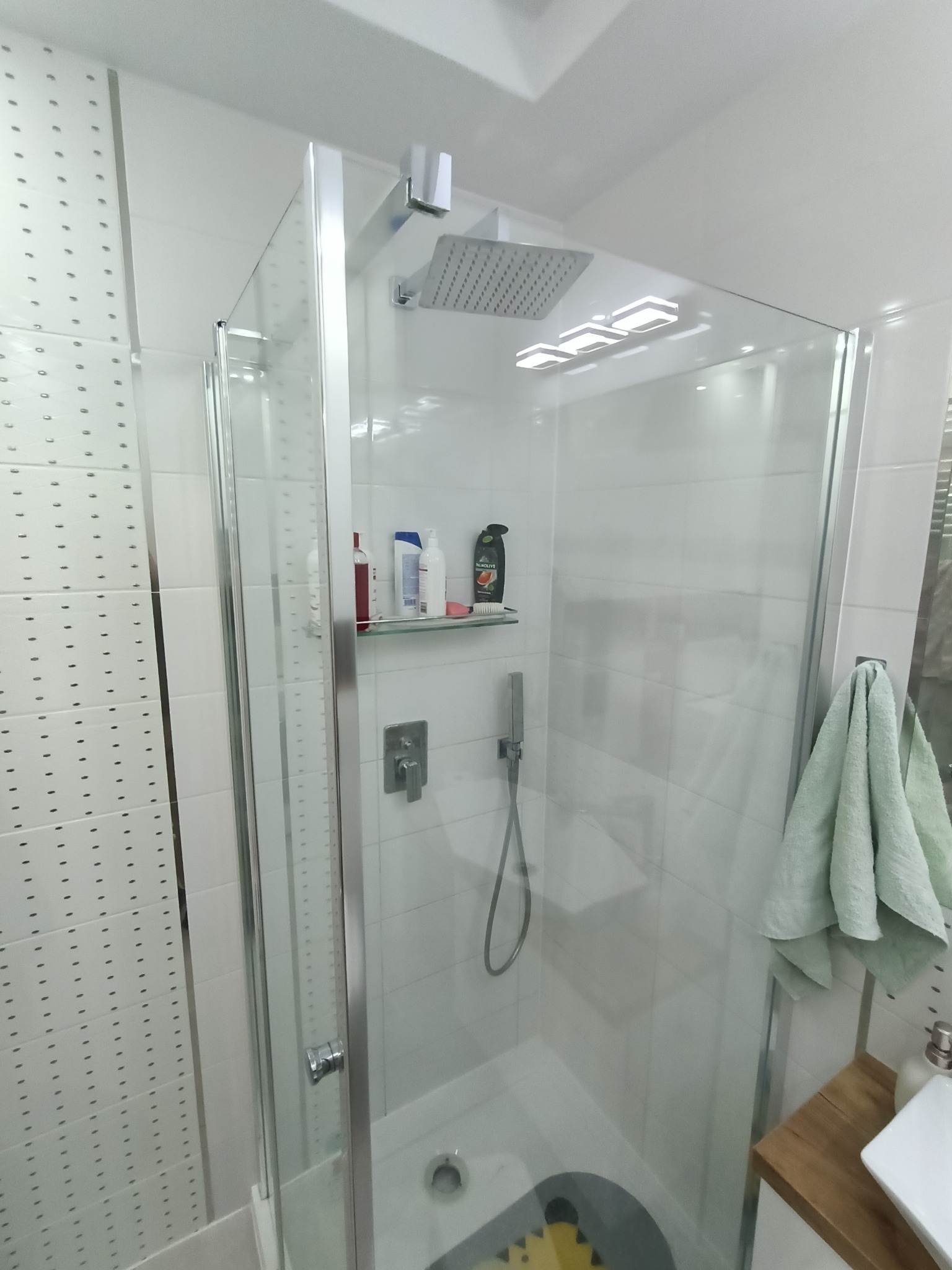 Nowoczesna kabina prysznicowa z deszczownicą i półką na kosmetyki, widok z boku, szara mata antypoślizgowa na dnie kabiny.