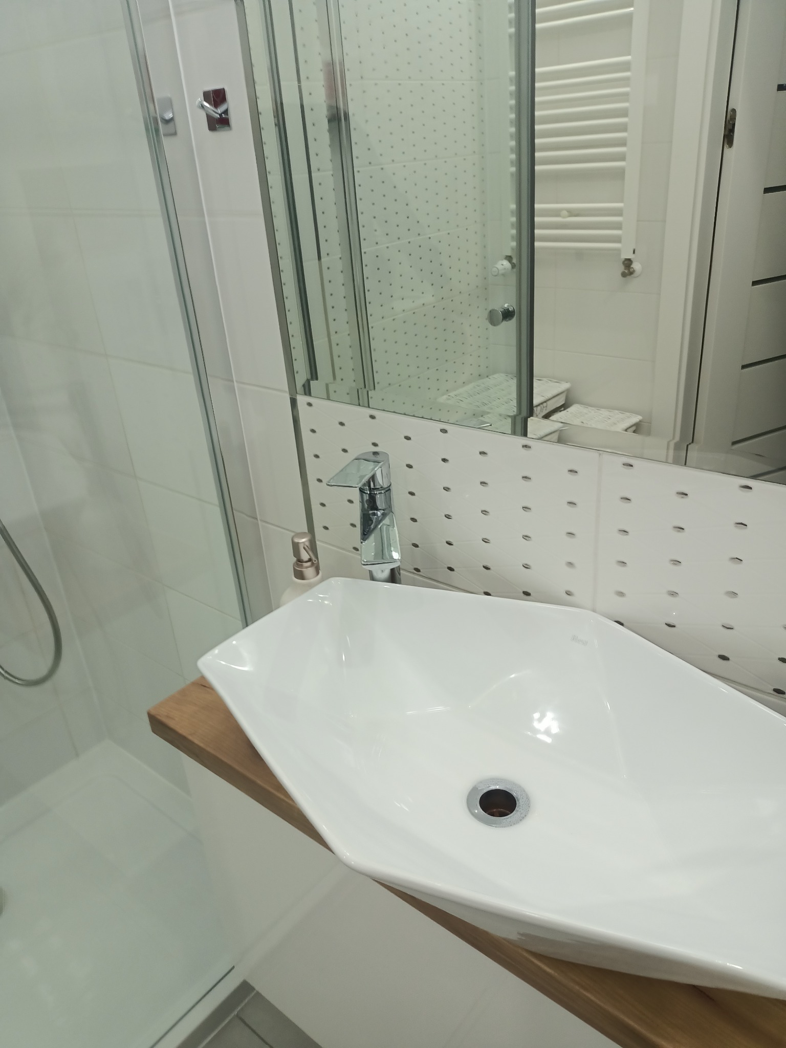 Nowoczesna łazienka z białą, asymetryczną umywalką nablatową na drewnianym blacie, chromowaną armaturą i kabiną prysznicową w tle.