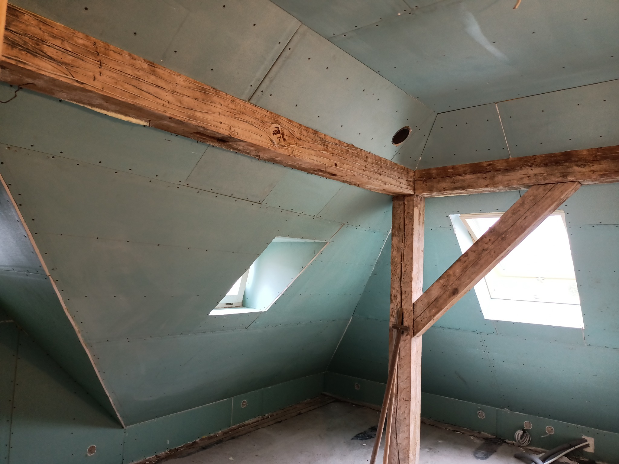 Poddasze z drewnianą konstrukcją, obłożone zielonymi płytami gipsowo-kartonowymi, widoczne okna dachowe i instalacje w podłodze.