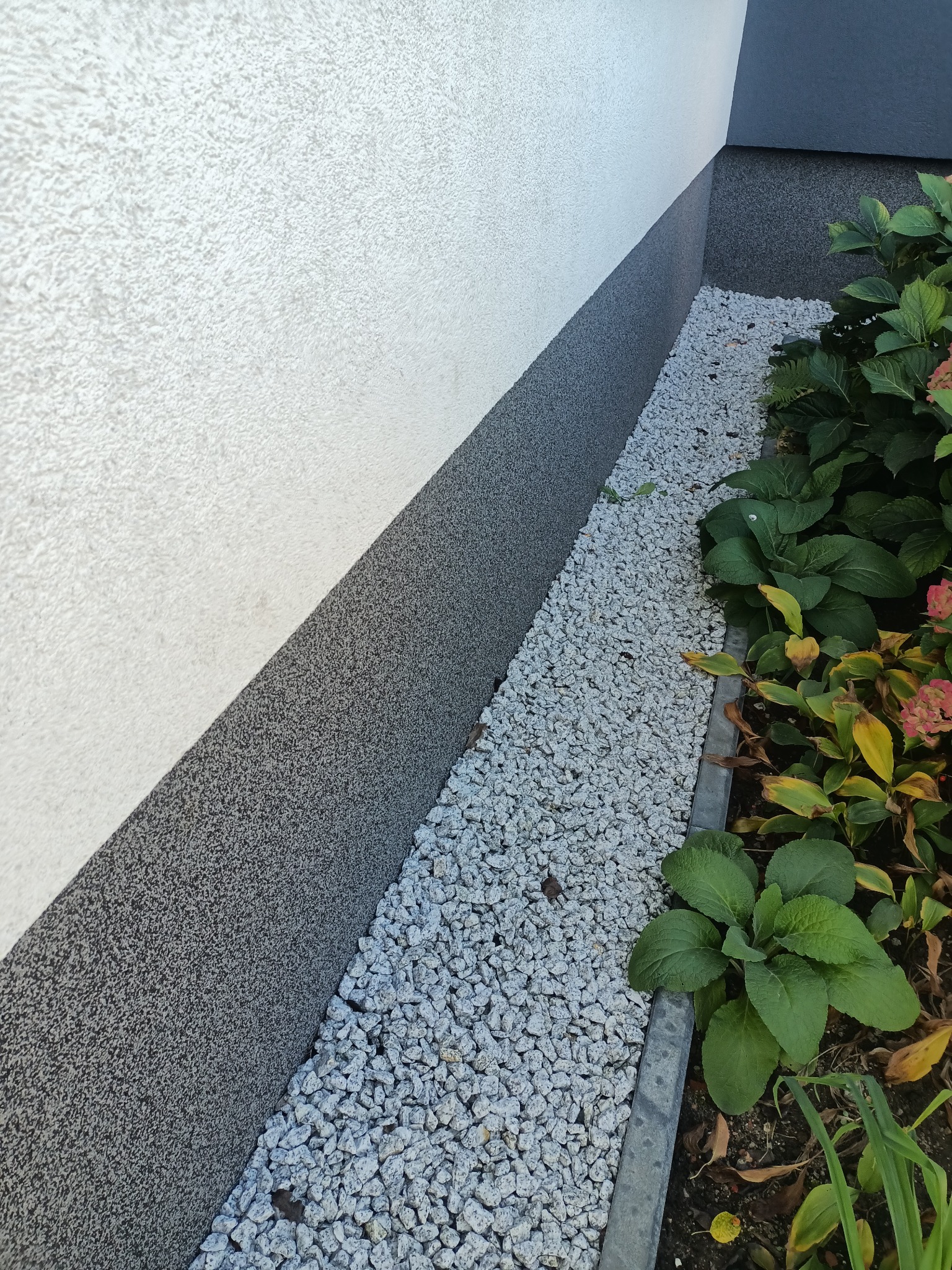 Dwuwarstwowa elewacja budynku z jasnoszarą górną częścią i ciemnoszarą dolną, oddzielona od rabaty z białym grysem betonowym obrzeżem.