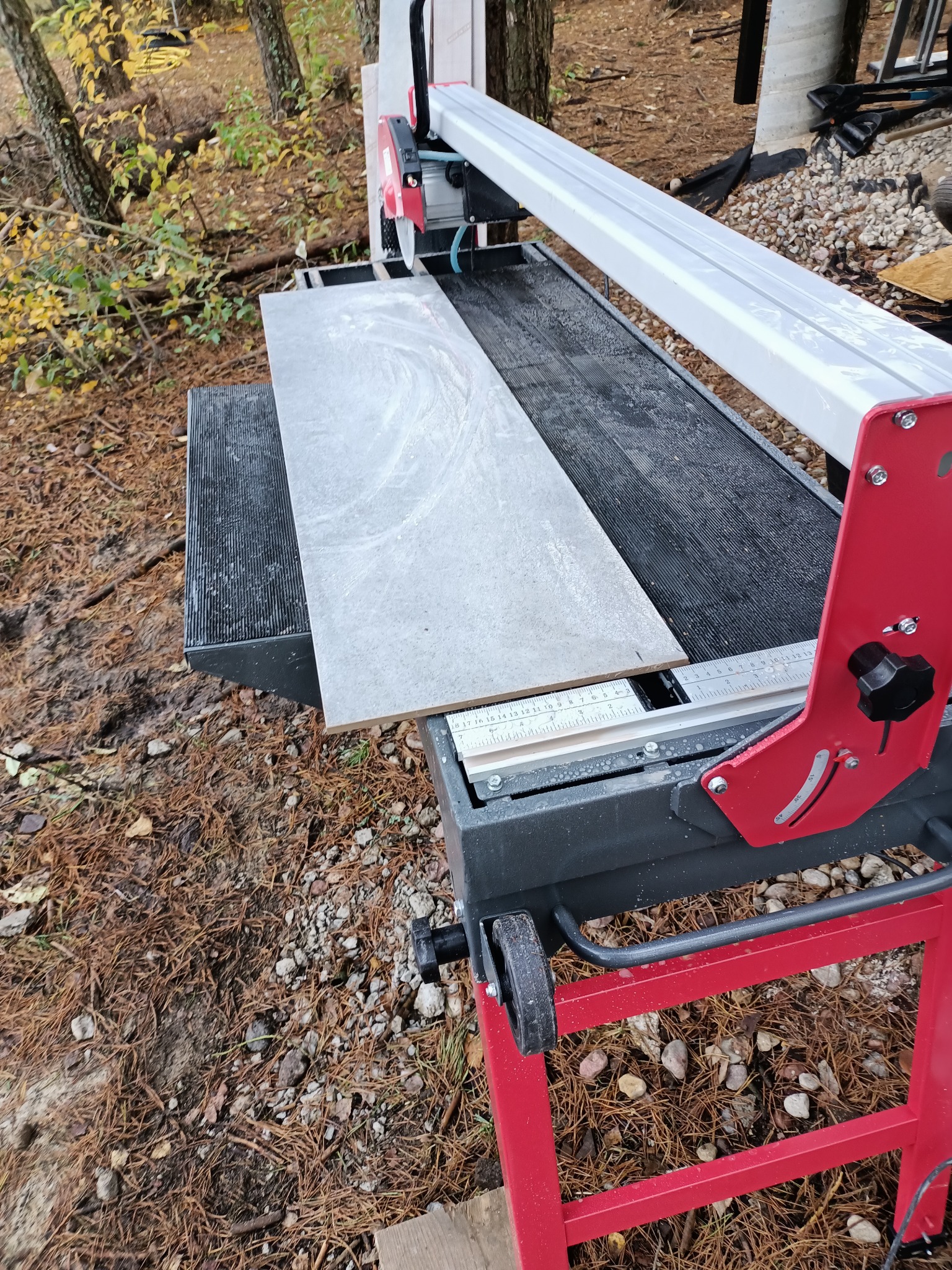 Szara płytka ceramiczna na stole z przecinarką do płytek z tarczą diamentową, widoczne otoczenie zewnętrzne z igliwiem.