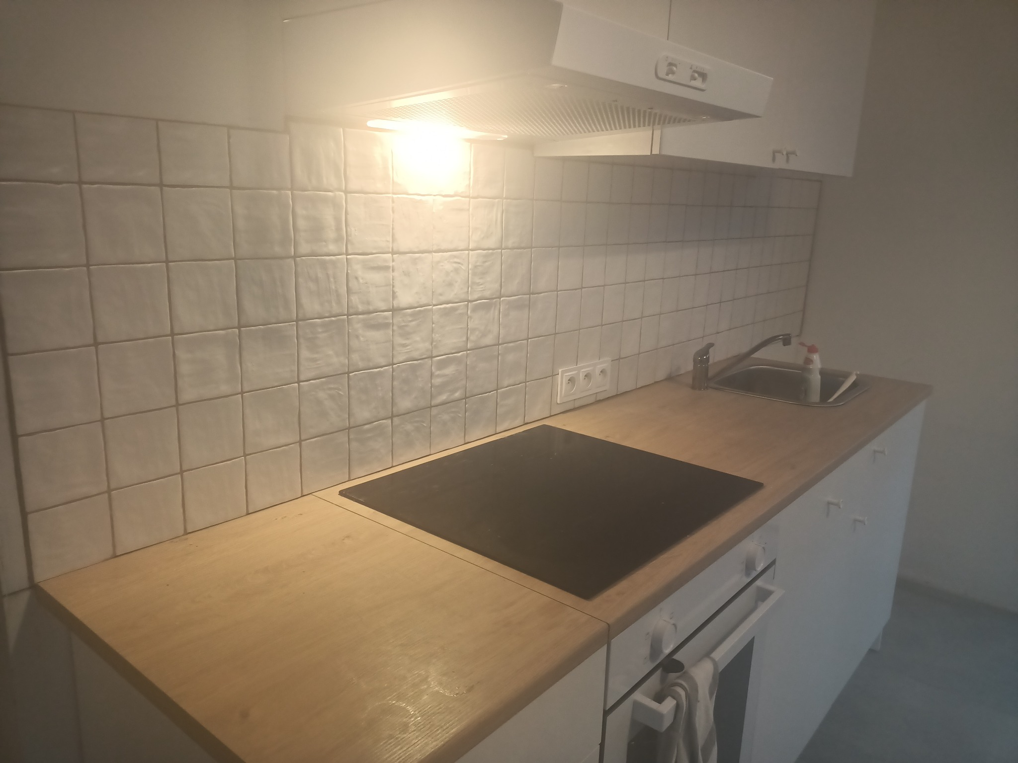 Wykończona kuchnia z białymi, kwadratowymi płytkami na ścianie, drewnianym blatem, czarną płytą grzewczą i białymi szafkami wiszącymi oraz piekarnikiem.