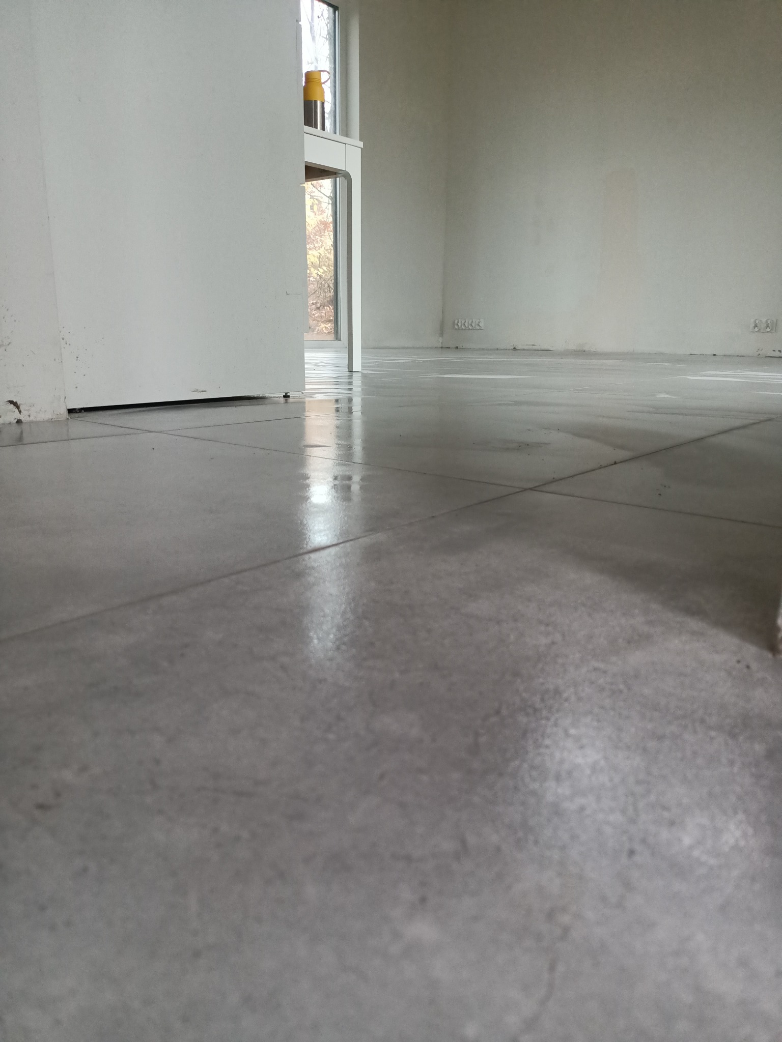 Nowo ułożona, błyszcząca, szara podłoga z płytek w pustym, jasnym pomieszczeniu. Widoczne fugi, ślady po pracy i gniazdka elektryczne na ścianie.