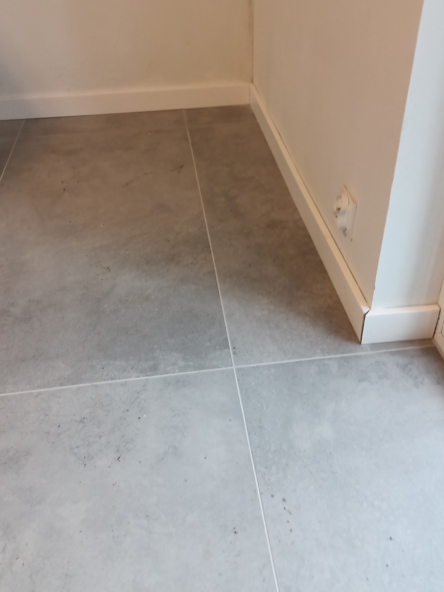 Szare płytki podłogowe z fugami i białą listwą przypodłogową oraz gniazdkiem elektrycznym na ścianie.