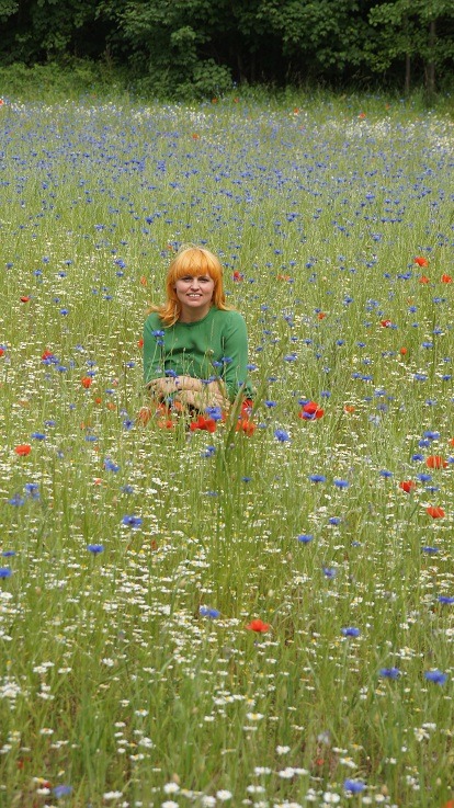 Kobieta z rudymi włosami w zielonej bluzce siedzi na łące pełnej chabrów, maków i rumianków.