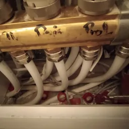 Rozdzielacz ogrzewania podłogowego z widocznymi rurami doprowadzającymi, opisany odręcznie markerem, w trakcie montażu, widoczne ślady instalacji.