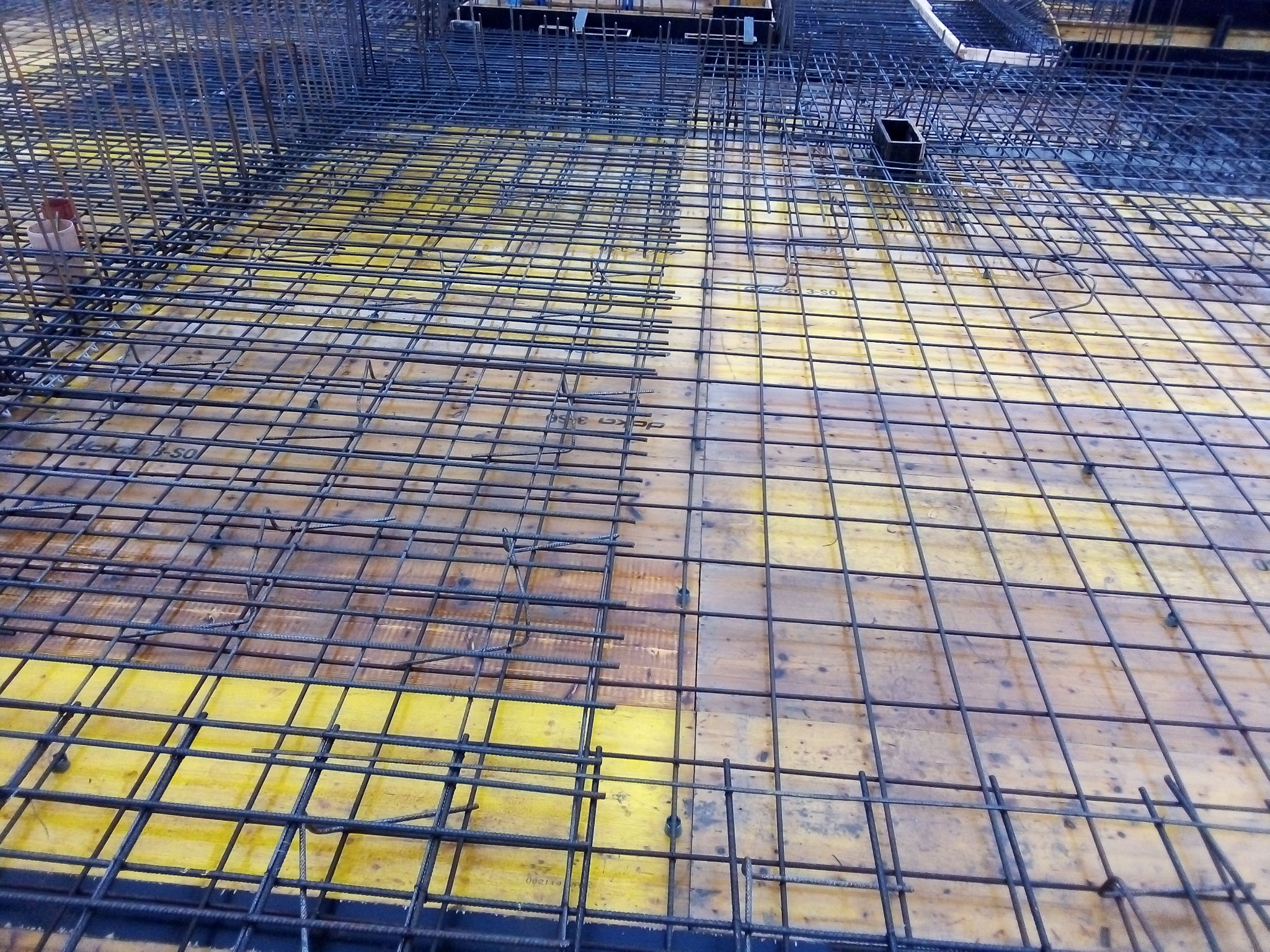 Zbrojenie płyty fundamentowej z widocznymi prętami stalowymi ułożonymi na deskowaniu z żółtej sklejki, przygotowane do zalania betonem.