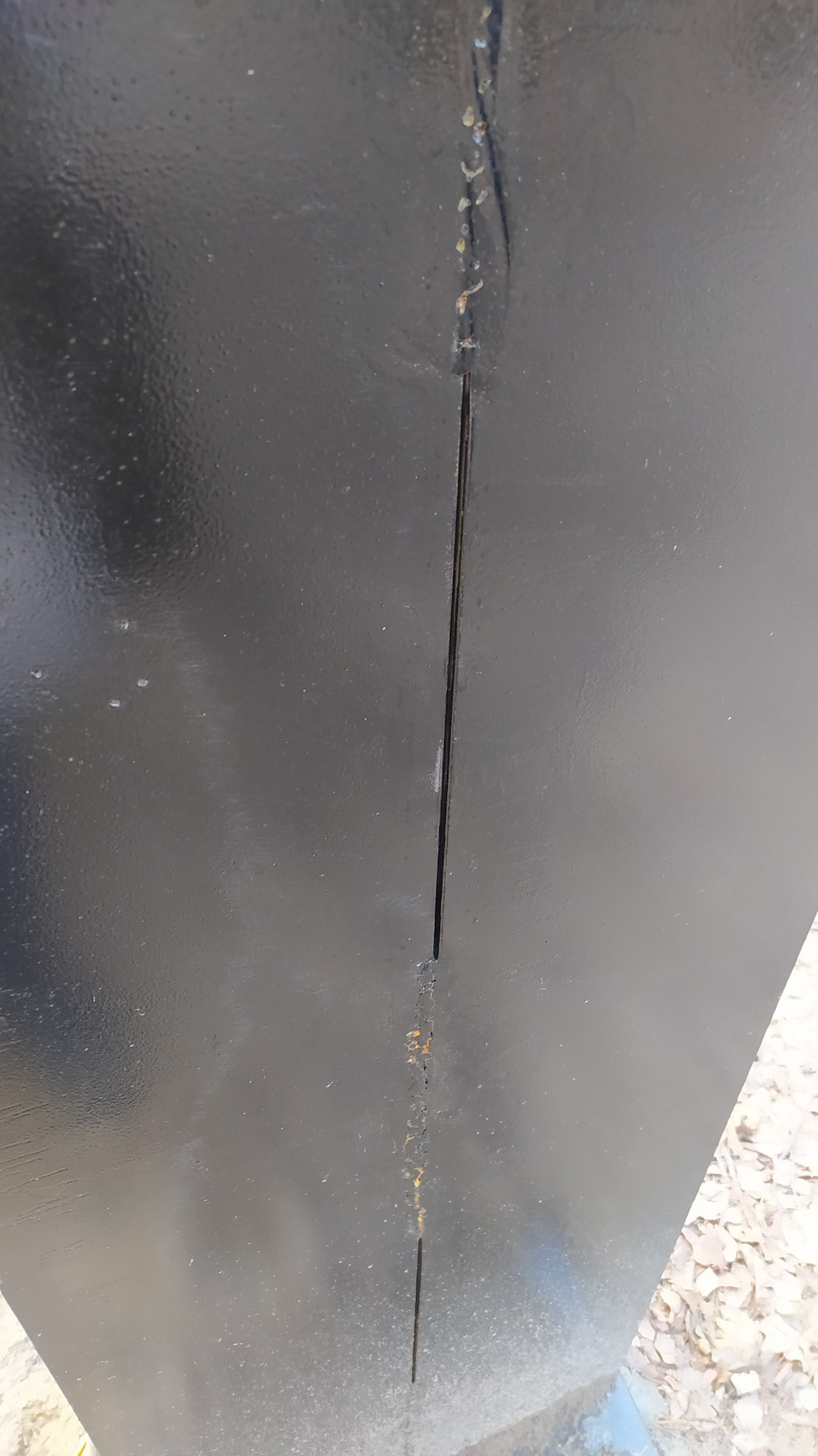 Fragment czarnej, metalowej konstrukcji z widocznym pionowym pęknięciem i śladami korozji wzdłuż niego.