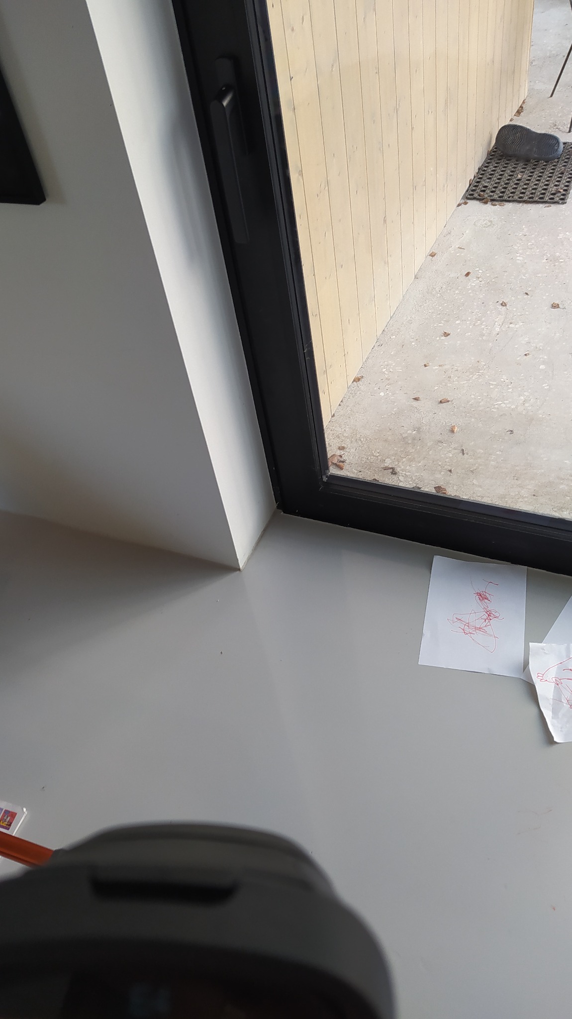 Fragment wnętrza z oknem w czarnej ramie, widok na zewnątrz z matą wejściową i butem, na parapecie leżą kartki z dziecięcymi rysunkami wykonanymi czerwonym długopisem.
