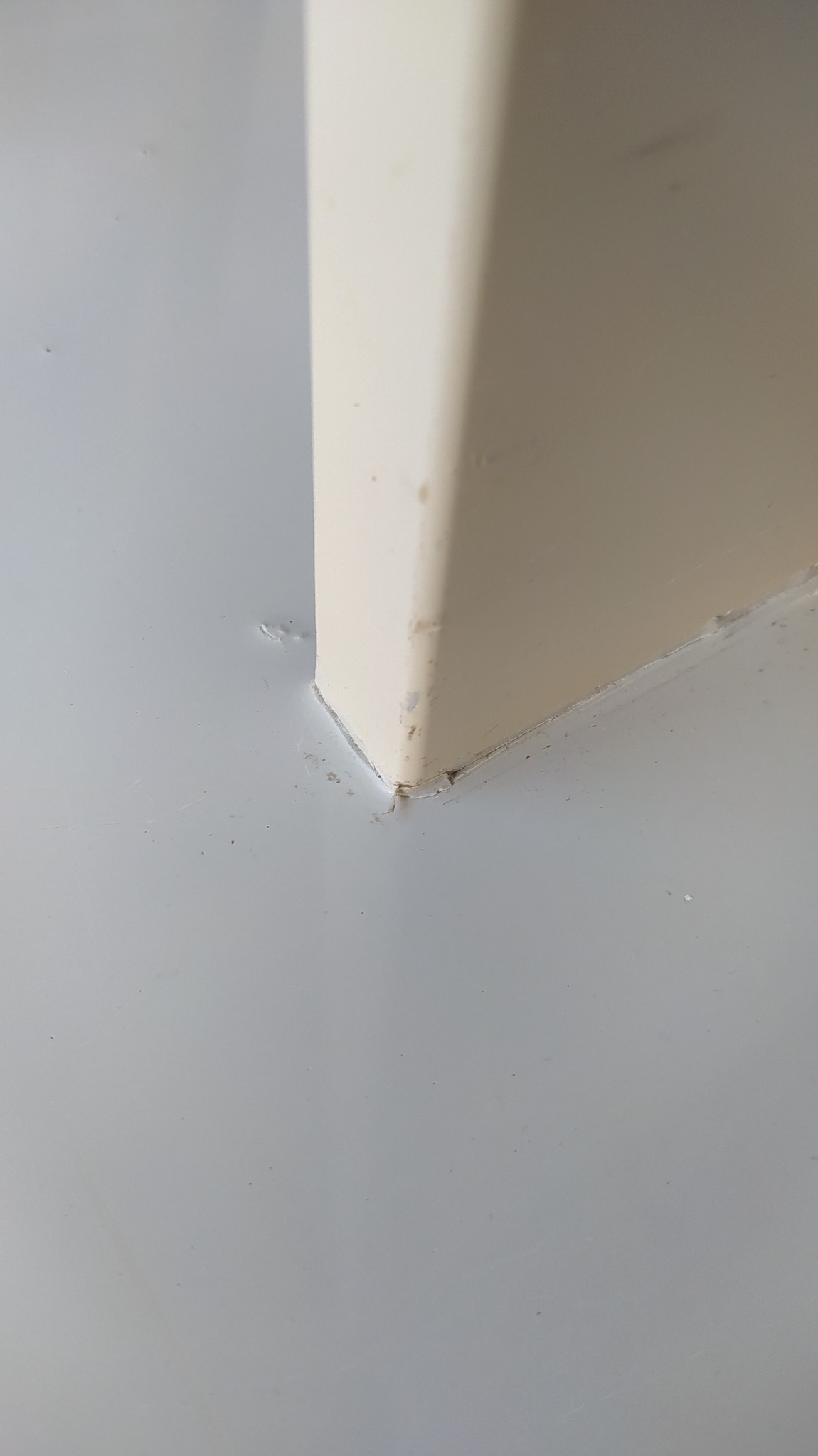 Dolny narożnik beżowej ściany przy szarej podłodze, widoczne niedoskonałości wykończenia, małe ubytki farby i silikonowe uszczelnienie. Niska jakość zdjęcia.