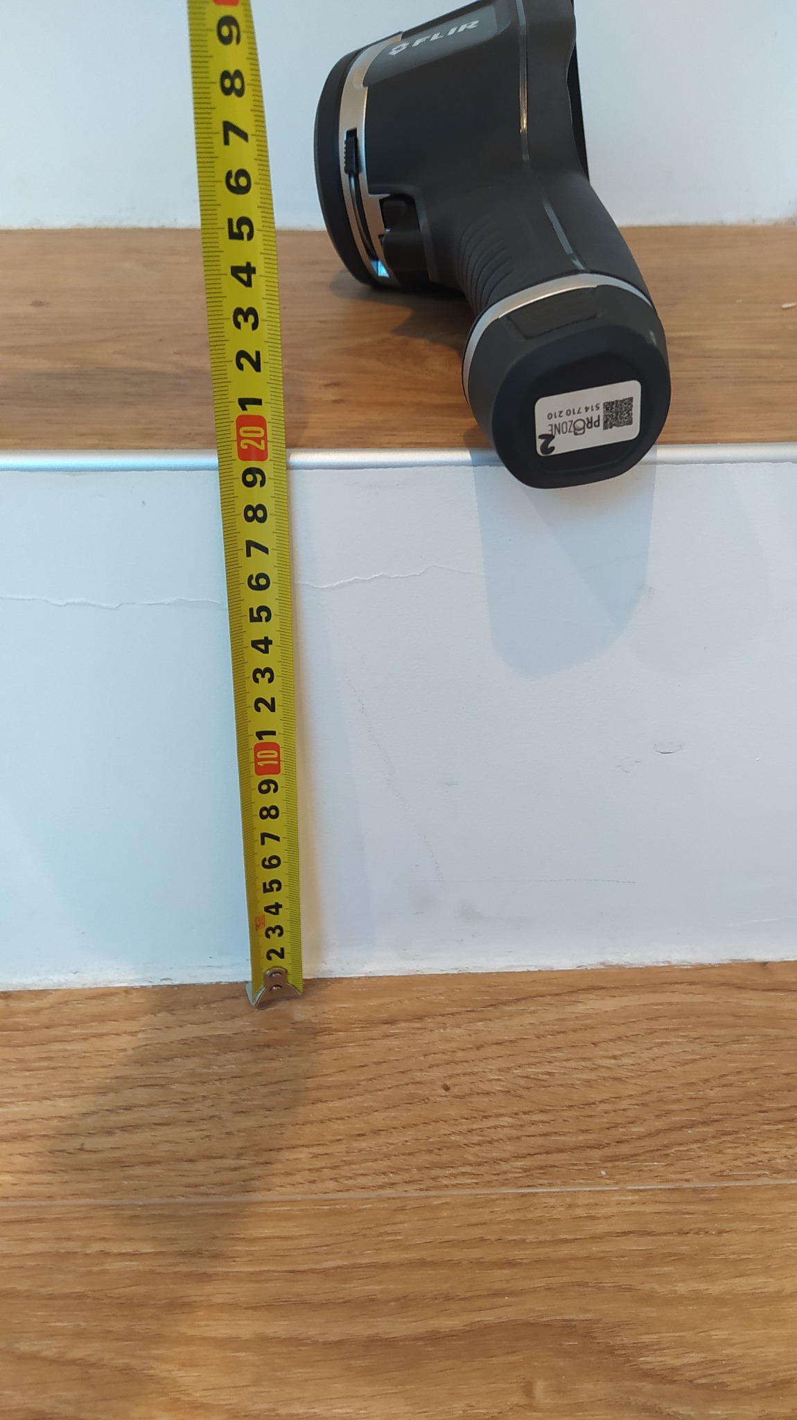 Pomiar termowizyjny przy użyciu kamery termowizyjnej FLIR, z widoczną miarką w centymetrach, umieszczoną na styku drewnianej podłogi i białej ściany.