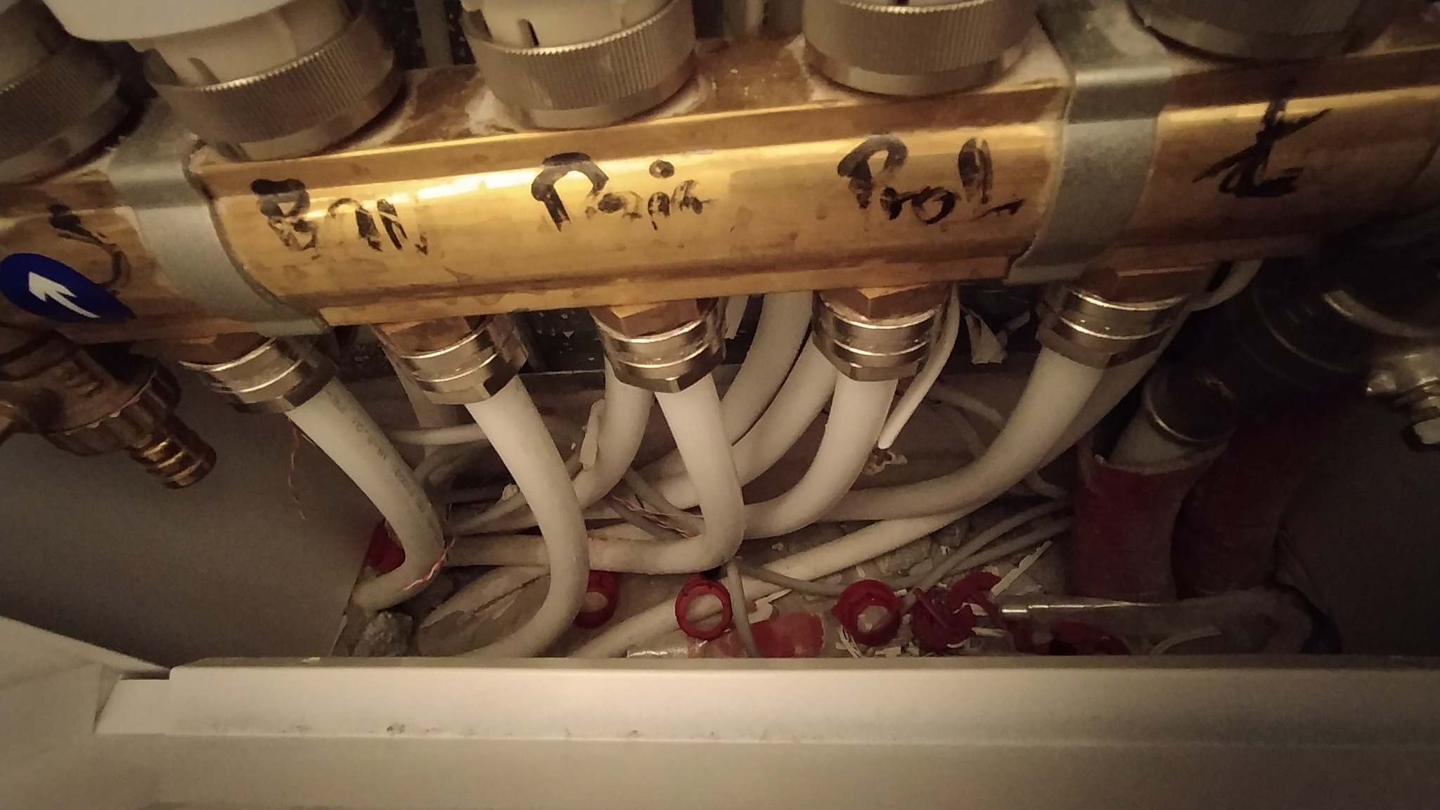 Rozdzielacz ogrzewania podłogowego z widocznymi rurami doprowadzającymi, opisany odręcznie markerem, w trakcie montażu, widoczne ślady instalacji.