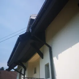 Fragment elewacji budynku z zamontowanym, ciemnym systemem orynnowania i elementami konstrukcyjnymi dachu, widzianymi z dołu pod kątem.