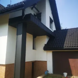 Dwukondygnacyjny dom z czarnym dachem z dachówki ceramicznej, czarnymi obróbkami blacharskimi i białymi ścianami, wykończony cegłą klinkierową na parterze, z widoczną anteną na dachu.