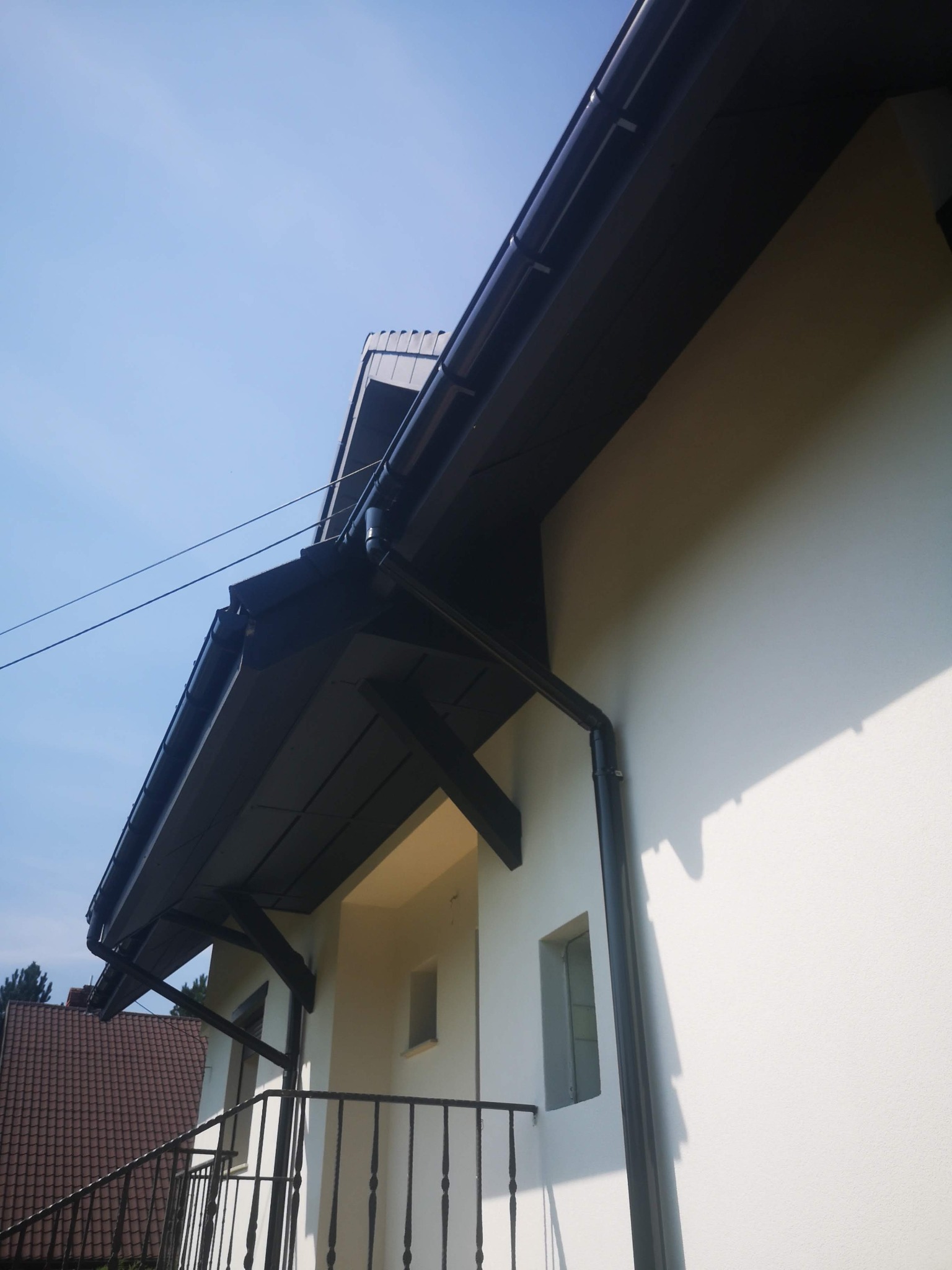 Fragment elewacji budynku z zamontowanym, ciemnym systemem orynnowania i elementami konstrukcyjnymi dachu, widzianymi z dołu pod kątem.