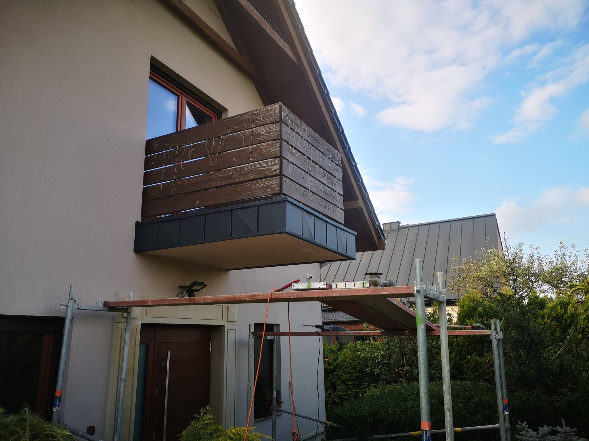 Dom z balkonem w trakcie montażu obróbek blacharskich, widoczne rusztowanie i narzędzia.