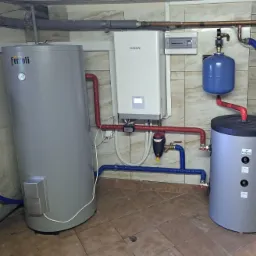 Szara pompa ciepła Ferroli obok białej pompy Kaisai, z widocznymi rurami w izolacji czerwonej i niebieskiej, oraz zbiornikiem wyrównawczym, na tle kafelkowanej ściany i podłogi.