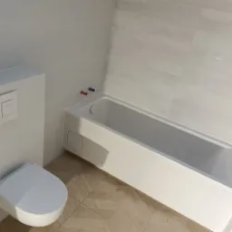 Minimalistyczna łazienka z białą zabudową WC i wanną, wykończona jasnymi płytkami imitującymi drewno na podłodze i szarymi, subtelnie wzorzystymi płytkami na ścianach.