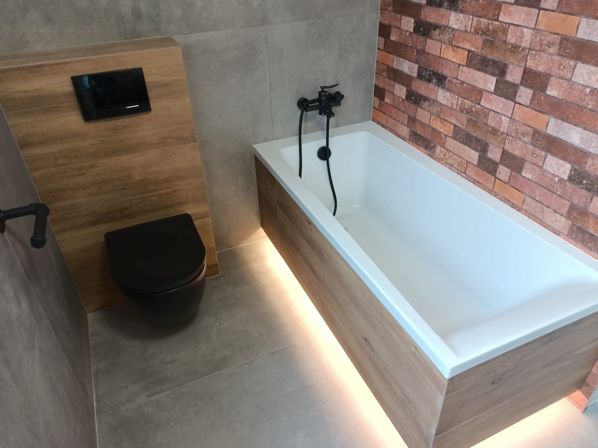 Nowoczesna łazienka z podświetlaną wanną obudowaną drewnem, czarną armaturą i podwieszaną toaletą w kolorze czarnym, wykończona płytkami w odcieniach szarości i brązową cegłą.
