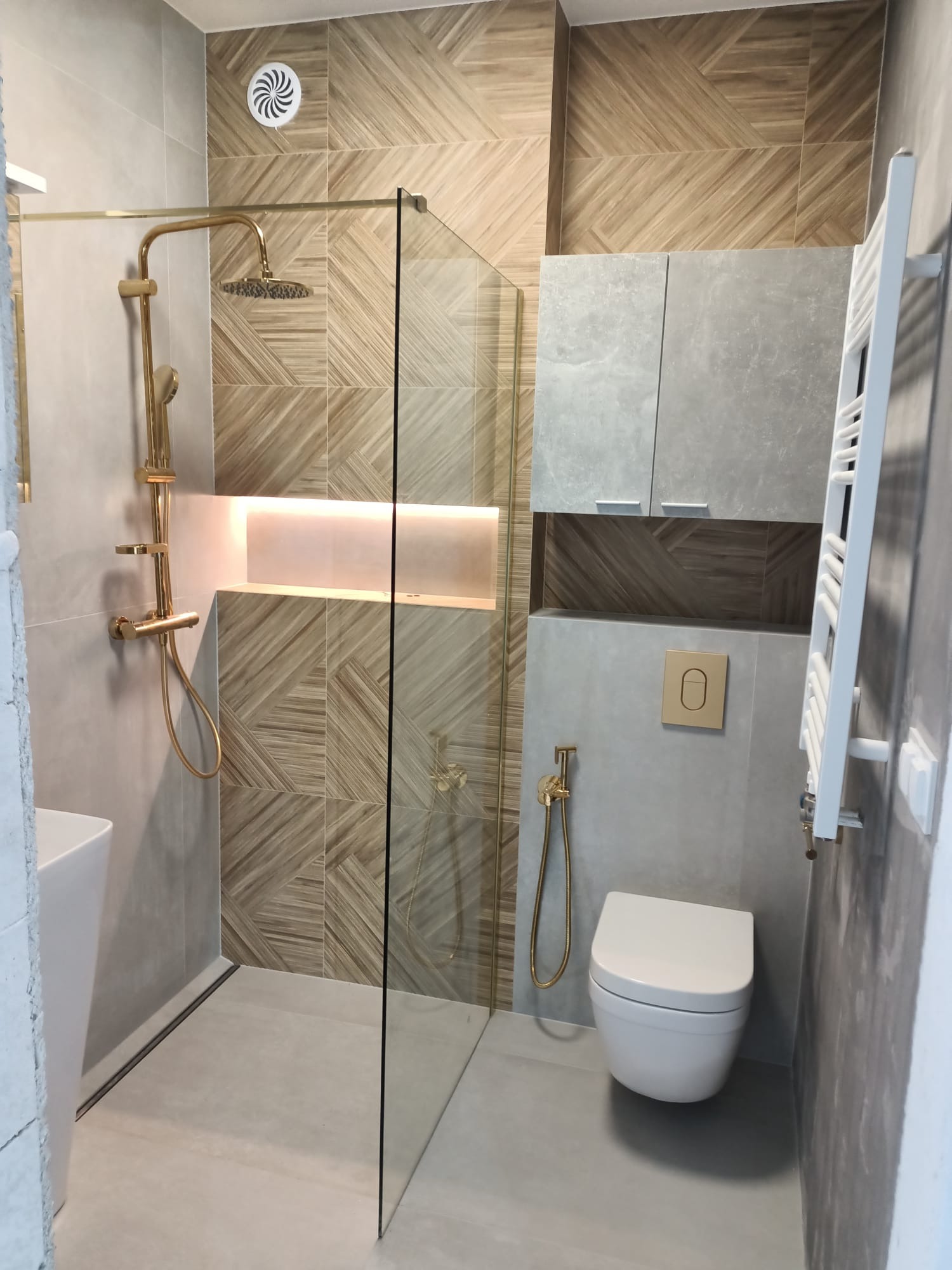 Nowoczesna łazienka z kabiną prysznicową typu walk-in, złotą armaturą i geometrycznymi płytkami na ścianach, oświetlona wnęka nad prysznicem.